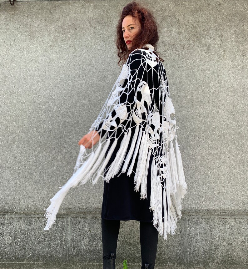Pu&ograve; includere: Scialle in crochet bianco con un design a forma di teschio e frange. Lo scialle &egrave; drappeggiato sulle spalle di una persona e viene indossato con un abito nero.