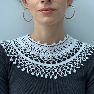 May include: A white crocheted collar with a delicate lace pattern. The collar is worn over a black shirt.
