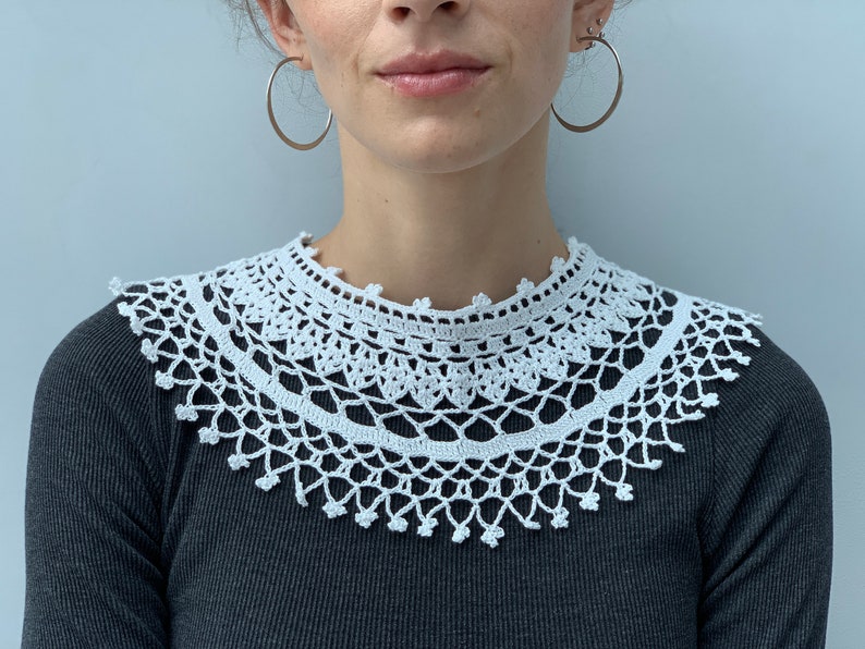 May include: A white crocheted collar with a delicate lace pattern. The collar is worn over a black shirt.