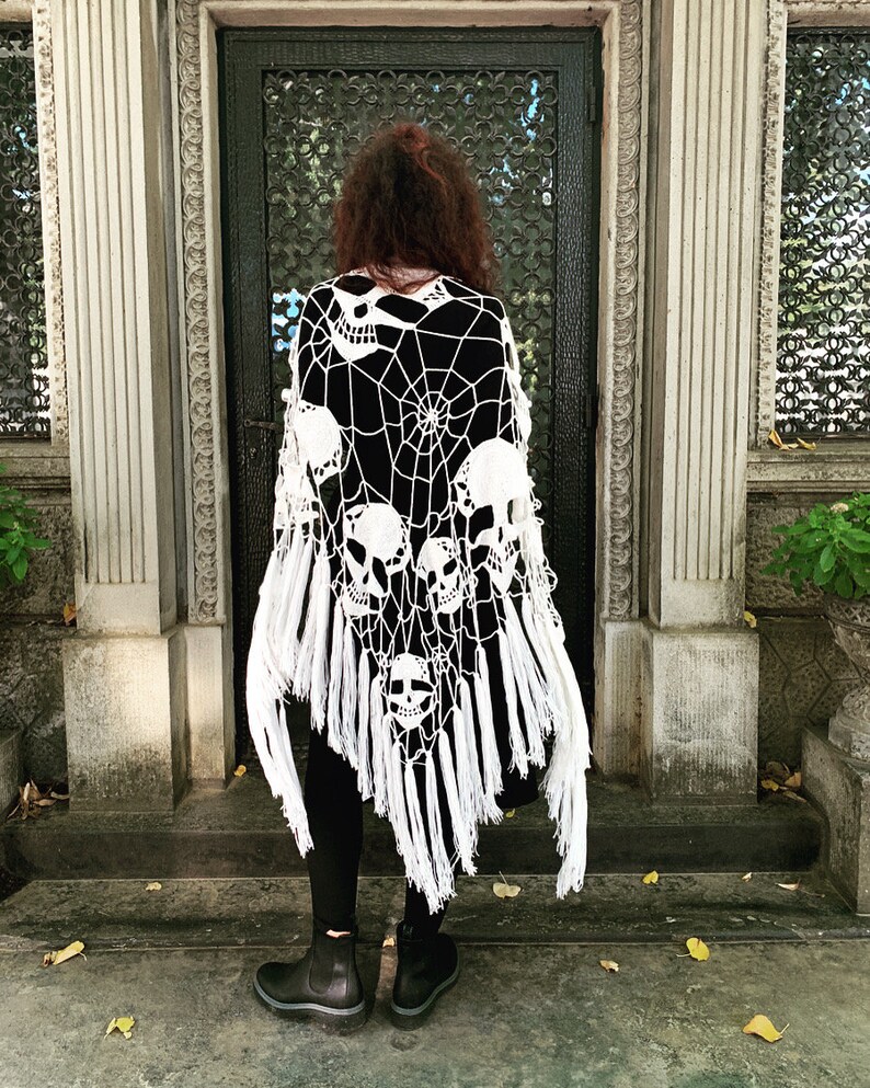 Pu&ograve; includere: Scialle in crochet bianco con un design a forma di teschio e ragnatela. Lo scialle ha frange lungo il bordo inferiore.