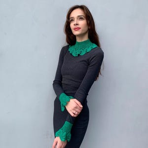 May include: A woman wearing a dark gray long-sleeved shirt with a green crocheted collar and matching cuffs.