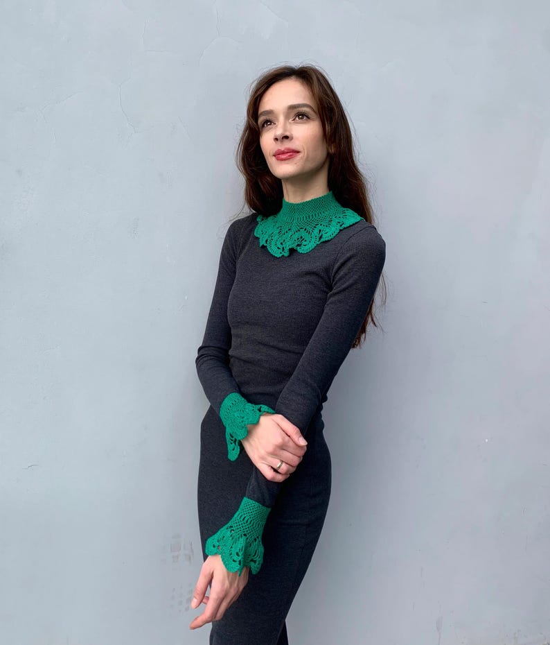 May include: A woman wearing a dark gray long-sleeved shirt with a green crocheted collar and matching cuffs.