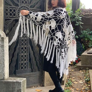 Pu&ograve; includere: Scialle in crochet bianco con un motivo a teschi e frange. Lo scialle &egrave; drappeggiato sulle spalle di una persona.