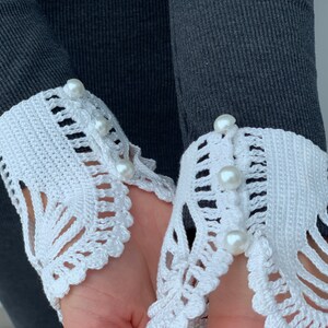 May include: A pair of white crocheted cuffs with pearl buttons. The cuffs are attached to a gray sweater.