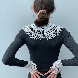 May include: A black long-sleeved shirt with a white crocheted collar and matching cuffs. The collar is wide and has a scalloped edge. The cuffs are also scalloped and have a pearl button detail.