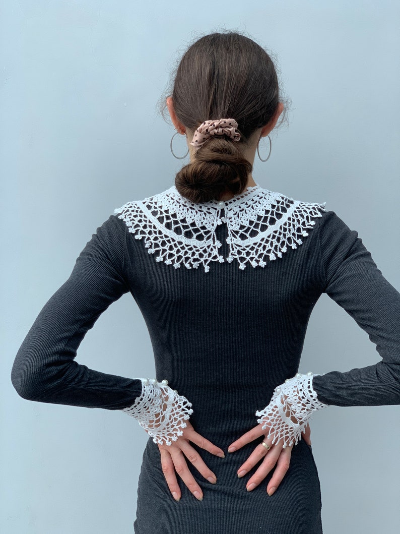 May include: A black long-sleeved shirt with a white crocheted collar and matching cuffs. The collar is wide and has a scalloped edge. The cuffs are also scalloped and have a pearl button detail.