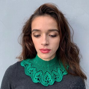 May include: A green crocheted collar with a delicate lace pattern. The collar is worn by a person with long brown hair.