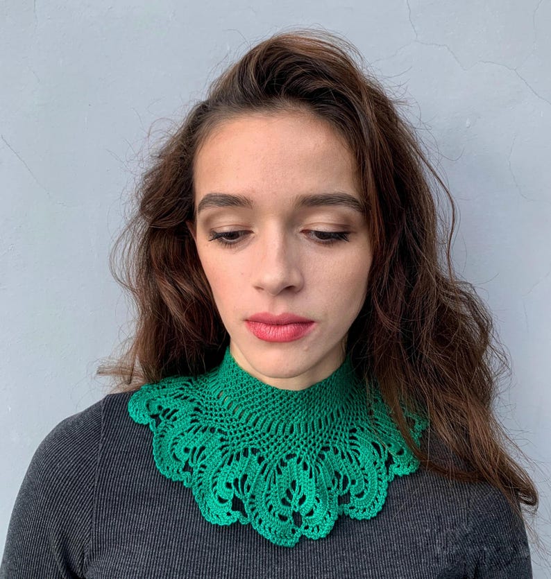 May include: A green crocheted collar with a delicate lace pattern. The collar is worn by a person with long brown hair.