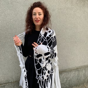 Pu&ograve; includere: Scialle bianco all'uncinetto con un design a forma di teschio e frange. Lo scialle &egrave; drappeggiato sulle spalle di una persona.