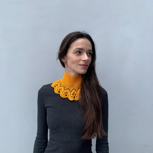 Puede incluir: Una mujer lleva un collar de ganchillo amarillo con un borde festoneado. Lleva una camisa de manga larga gris oscuro.