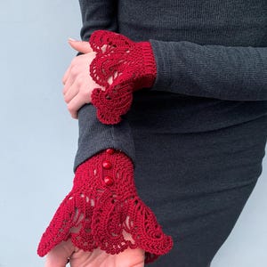 May include: Red crocheted lace cuffs with buttons, worn on a dark gray long-sleeved shirt.