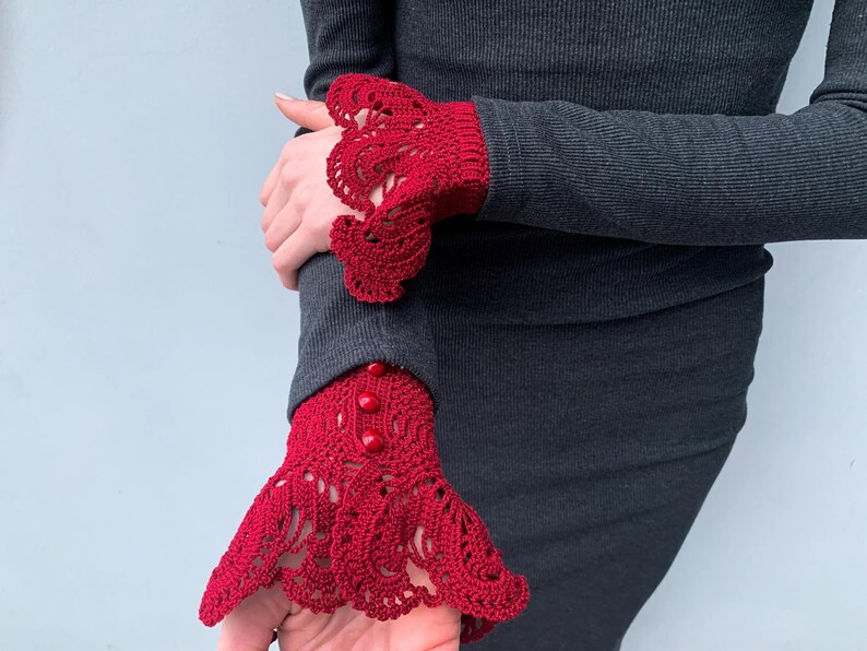 May include: Red crocheted lace cuffs with buttons, worn on a dark gray long-sleeved shirt.