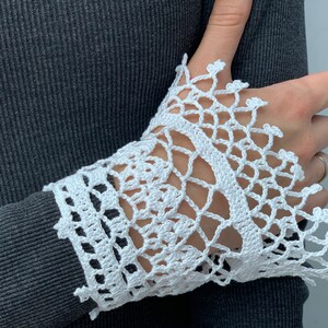 May include: White crocheted lace cuff with a delicate pattern. The lace is attached to a gray sweater sleeve.