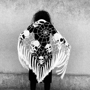 Pu&ograve; includere: Un scialle bianco all'uncinetto con un disegno a ragnatela e motivi di teschi. Lo scialle ha frange lunghe ed &egrave; drappeggiato sulle spalle di una persona.