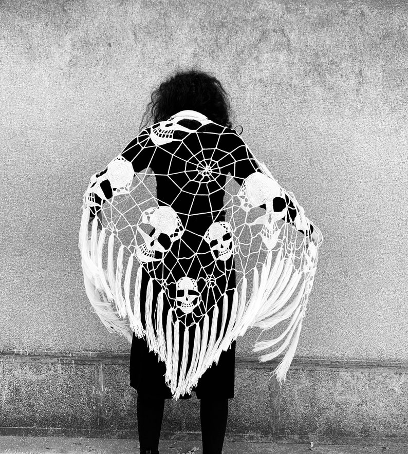 Pu&ograve; includere: Un scialle bianco all'uncinetto con un disegno a ragnatela e motivi di teschi. Lo scialle ha frange lunghe ed &egrave; drappeggiato sulle spalle di una persona.