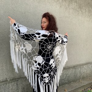 Pu&ograve; includere: Scialle in crochet bianco con un design a forma di teschio e ragnatela. Lo scialle ha lunghe frange bianche.