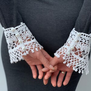 May include: White crochet lace cuffs with pearl embellishments. The cuffs are attached to a black long-sleeved shirt.