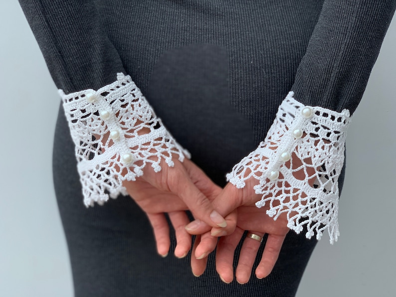 May include: White crochet lace cuffs with pearl embellishments. The cuffs are attached to a black long-sleeved shirt.