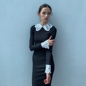 May include: A woman wearing a black long-sleeved dress with a white lace collar and cuffs. The dress has a fitted silhouette and the lace details are delicate and feminine.