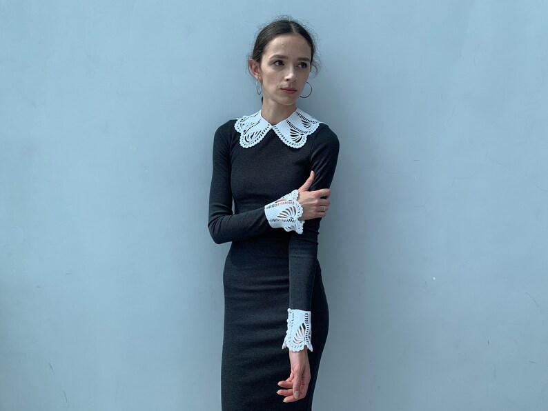 May include: A woman wearing a black long-sleeved dress with a white lace collar and cuffs. The dress has a fitted silhouette and the lace details are delicate and feminine.