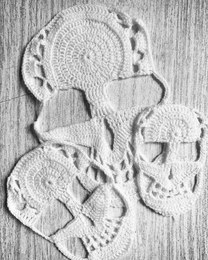 Pu&ograve; includere: Tre forme di teschio in crochet bianco, con dettagli intricati, sono disposte su una superficie in legno chiaro.