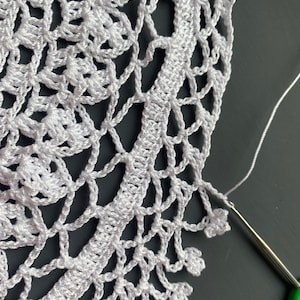 May include: Close-up of a white crocheted doily with a green crochet hook and white yarn. The doily has a delicate floral pattern and a scalloped edge.