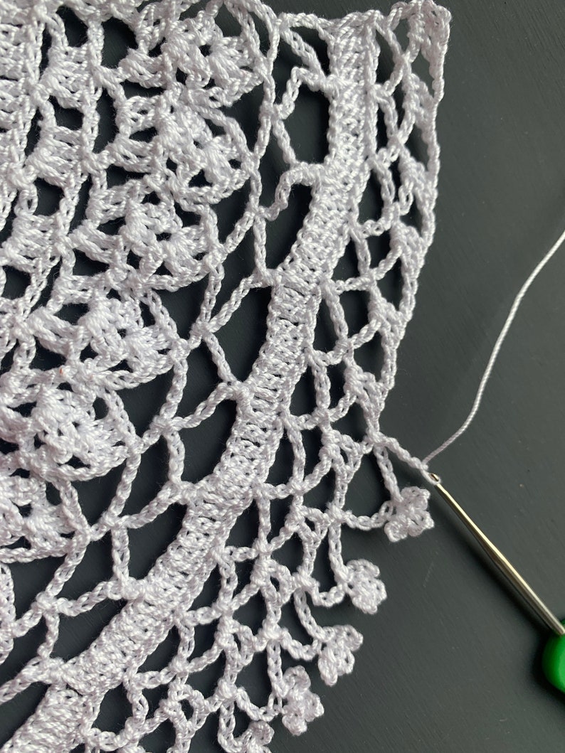 May include: Close-up of a white crocheted doily with a green crochet hook and white yarn. The doily has a delicate floral pattern and a scalloped edge.