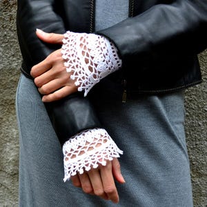 May include: White, open-weave crochet cuffs worn with a black leather jacket. The cuffs have a scalloped edge and are worn over a gray dress. The jacket has a zipper closure.