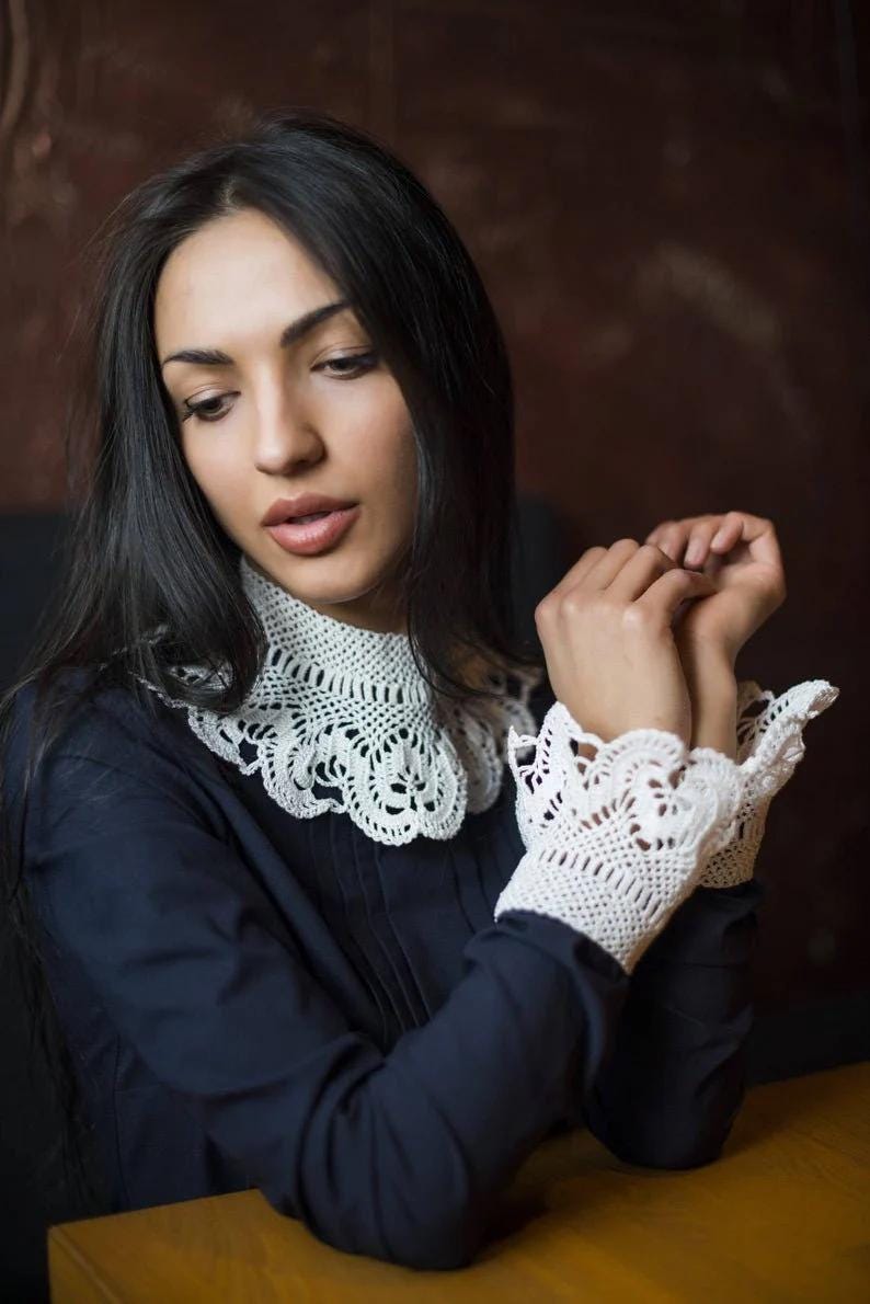 May include: A woman wearing a navy blue blouse with a white crocheted collar and matching cuffs. The collar and cuffs have a delicate floral design.