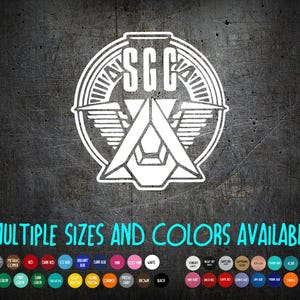 May include: White SGC logo with a stylized design featuring wings and geometric shapes, set against a dark, textured background. The text "MULTIPLE SIZES AND COLORS AVAILABLE" is displayed below the logo, with a color palette.