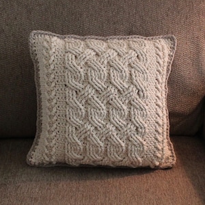 Puede incluir: Una almohada blanca de crochet con un patrón de punto de cable sobre un sillón marrón.