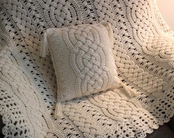 Peut inclure: Une couverture en crochet blanc avec un motif en tricot torsadé et un coussin assorti. La couverture est drapé sur un fauteuil marron.