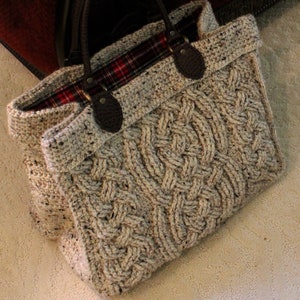 May include: A large, crocheted tote bag with a brown leather handle and a plaid lining. The bag is made with a cable stitch pattern and has a textured, rustic look.