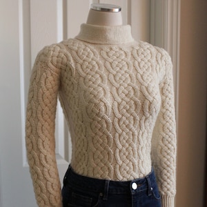 Op de afbeelding: Een crèmekleurige kabelgebreide trui met een rolkraag. De trui is op een mannequin en heeft een gedetailleerd kabelgebreid patroon.