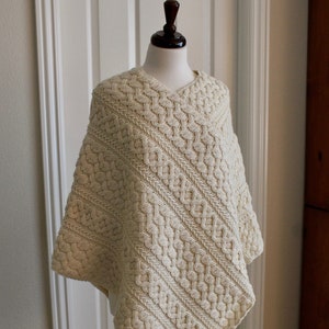 Puede incluir: Un poncho de punto de color crema con un patrón de punto de cable. El poncho se muestra en un maniquí.