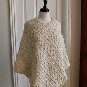 Può includere: Un poncho di maglia color crema con un motivo a treccia. Il poncho è esposto su un manichino.