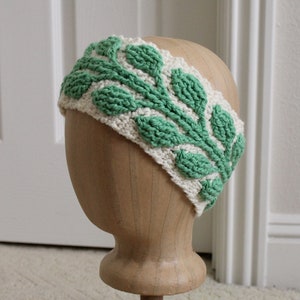 May include: A cream-colored, crocheted headband with a green leaf pattern. The leaves are raised and textured, creating a three-dimensional effect. The headband is displayed on a wooden head form.