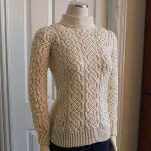 May include: A cream-coloured cable knit sweater with a high neck and long sleeves.