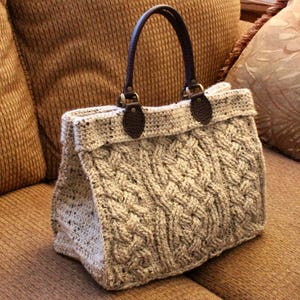 May include: A beige crocheted tote bag with brown leather handles and hardware. The bag has a cable knit pattern and a textured finish.