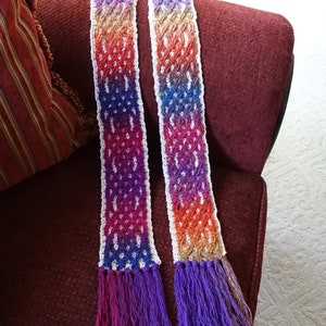 May include: A handmade, colourful scarf with a gradient pattern. The scarf transitions from purple to orange, with white accents and a fringe at the ends. The scarf is approximately 152 cm long.