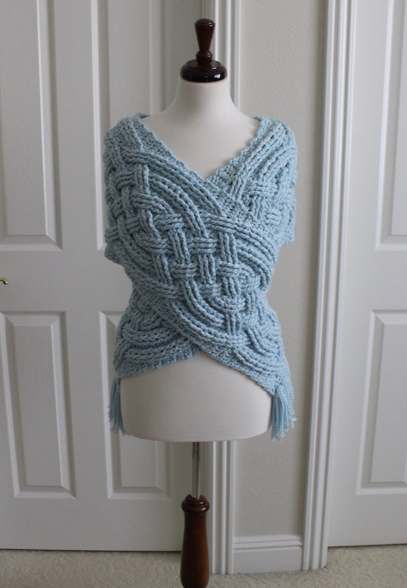 May include: A light blue crocheted shawl with a braided pattern and fringe. The shawl is displayed on a white mannequin.