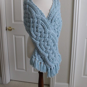 May include: A light blue knitted shawl with a cable knit pattern and fringe. The shawl is draped over a wooden mannequin.