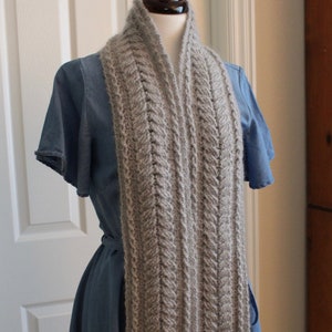 Puede incluir: Una bufanda de punto gris con un patrón de trenza. La bufanda está drapeada sobre un maniquí que lleva un vestido azul.