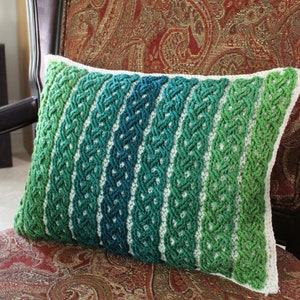 Peut inclure: Un coussin crocheté vert, bleu et blanc avec un motif tressé. Le coussin est sur un fauteuil à motifs marrons et dorés.