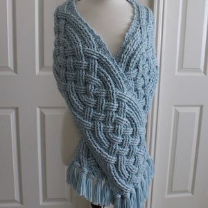 May include: A light blue crocheted shawl with a cable knit pattern and tassels. The shawl is draped over a mannequin.