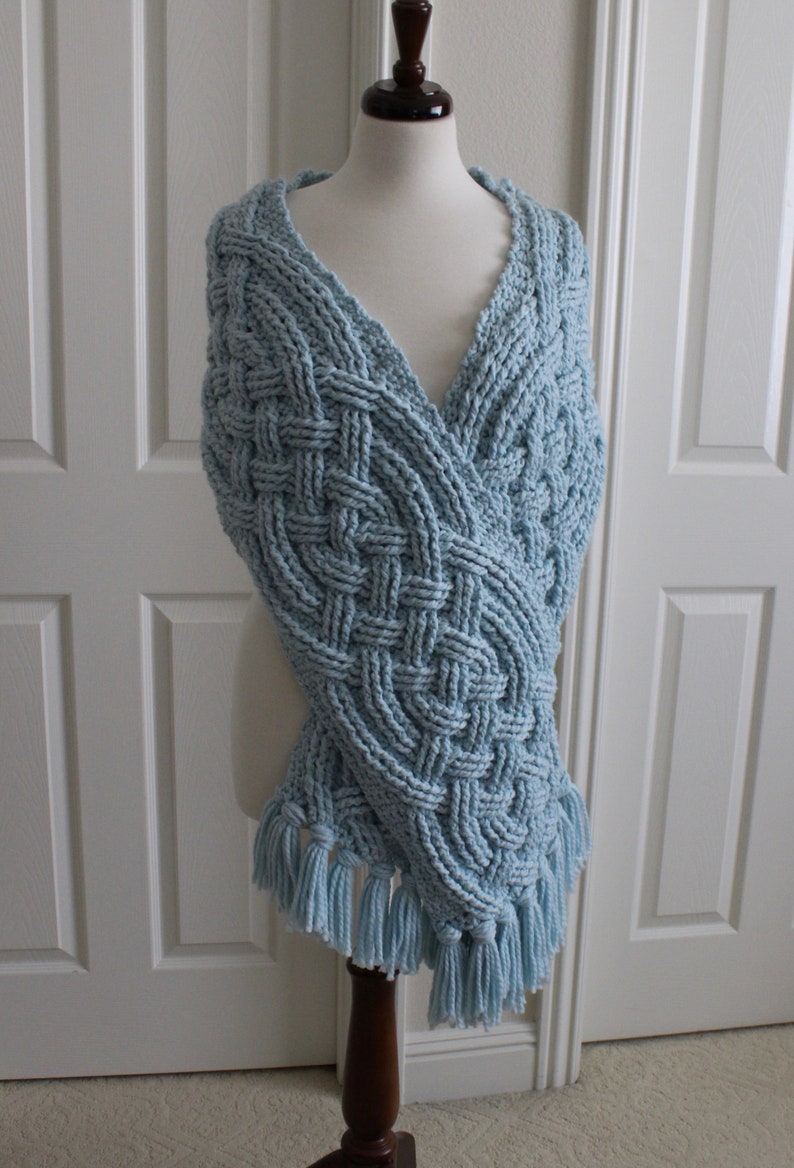 May include: A light blue crocheted shawl with a cable knit pattern and tassels. The shawl is draped over a mannequin.