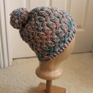 May include: A blue and pink knitted hat with a pom-pom on top. The hat is on a wooden display stand.