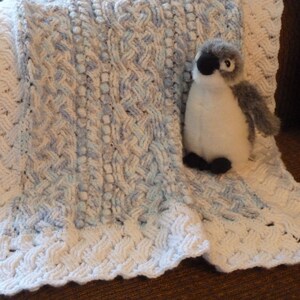 Peut inclure: Une couverture pour bébé en crochet blanc et bleu avec une bordure en crochet blanc. Un petit jouet en peluche en forme de pingouin est posé sur la couverture.