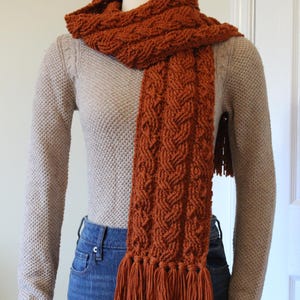 May include: A burnt orange cable-knit scarf with a fringed edge, displayed on a mannequin. The scarf is draped over a beige sweater and blue jeans. The knitted pattern and warm colour are the focal points, suggesting a cosy, handmade accessory.