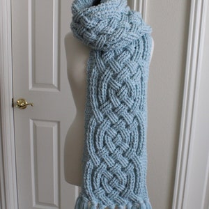 May include: A light blue knitted scarf with a braided pattern and tassels. The scarf is displayed on a white mannequin.